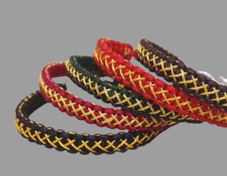K&ouml;nnte beinhalten: Vier geflochtene Armb&auml;nder mit einem Kreuzmuster. Die Armb&auml;nder sind schwarz, rot, gr&uuml;n und braun mit goldfarbenem Garn, das in das Design eingewebt ist.