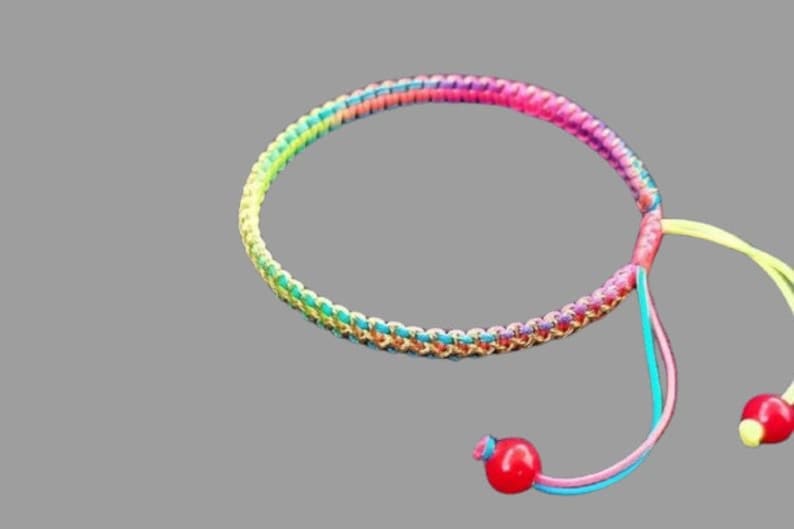 K&ouml;nnte beinhalten: Ein farbenfrohes geflochtenes Armband mit einem Regenbogenmuster und zwei roten Perlen an den Enden. Das Armband ist aus Schnur gefertigt und hat ein schlichtes Design.