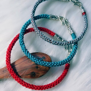 Może przedstawiać: Trzy plecione bransoletki ze sznurka z srebrnymi zapięciami. Bransoletki są czerwone, niebieskie i szare.