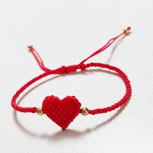 May include: A red woven bracelet featuring a heart-shaped charm. The bracelet has a braided design with gold-colored beads on either side of the heart. The adjustable cord is red.