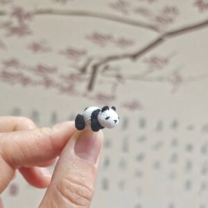 May include: A small crocheted panda bear figurine, lying on its side with its black and white fur details visible. The panda is resting on a finger.