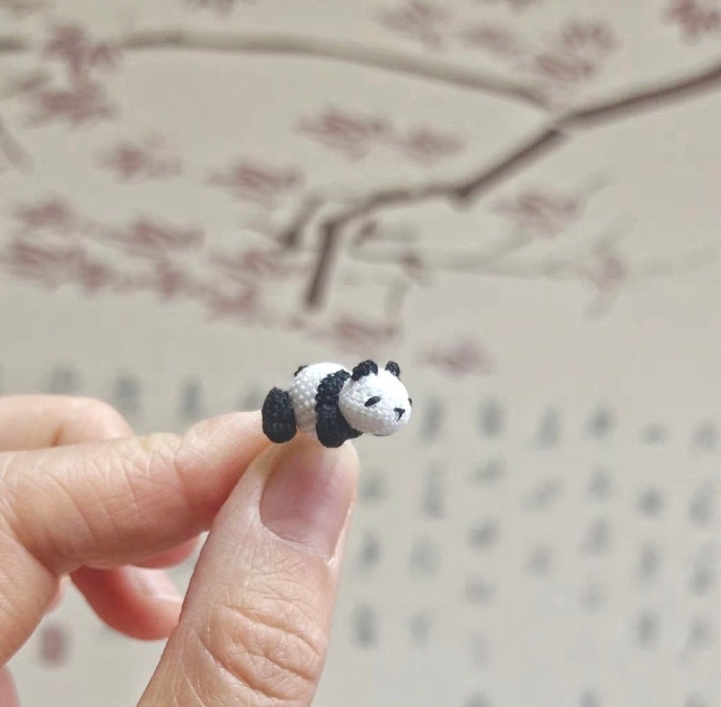 May include: A small crocheted panda bear figurine, lying on its side with its black and white fur details visible. The panda is resting on a finger.