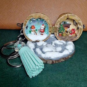 May include: A Christmas-themed walnut shell ornament with a snowman and Santa Claus scene on one side and a fireplace and Christmas tree scene on the other side. The ornament is attached to a silver key chain with a light blue tassel.
