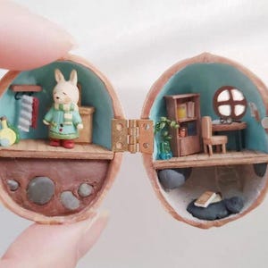 May include: A miniature diorama inside a hinged walnut shell featuring two levels. The left side shows a rabbit figurine in a green coat and scarf. The right side contains a wooden bookshelf, a desk with a chair, and a small round window.