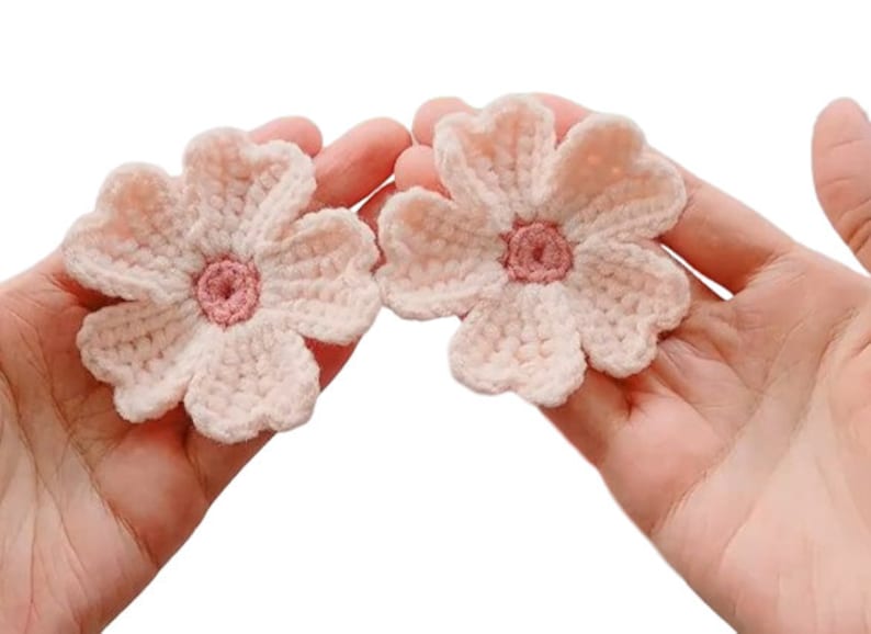 May include: Two crocheted pink flowers with a smaller pink center. The flowers are held in a person's hands.