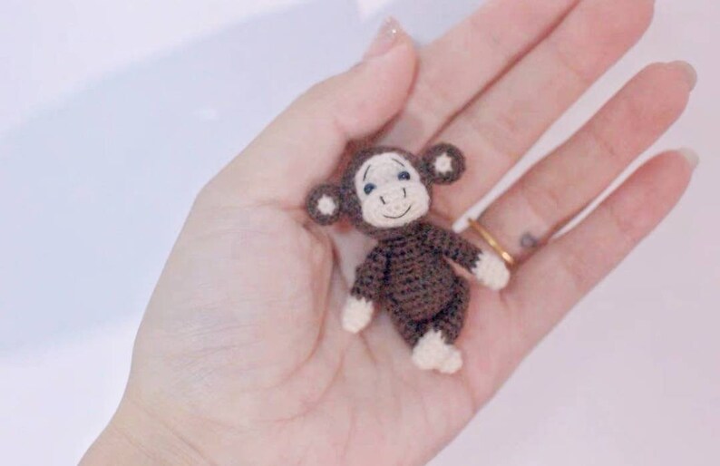 Puede incluir: Un peque&ntilde;o mono de crochet con pelaje marr&oacute;n, rasgos faciales blancos y ojos azules. El mono est&aacute; sentado en la palma de una mano.