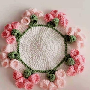 May include: A handmade crochet coaster featuring a central white circle with a spiral pattern. The coaster is adorned with pink and green crochet flowers around the edge, creating a floral design. The coaster is a decorative item.