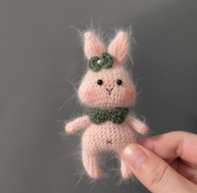 May include: A small pink crocheted bunny with green felt ears and a green felt bow. The bunny has black eyes and a pink nose.
