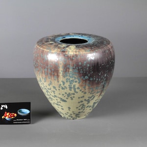 Puede incluir: Un jarrón de cerámica con un cuerpo ancho y redondeado y una abertura estrecha. El jarrón presenta un patrón moteado en tonos marrones, verdes y beige, con un borde azul. El jarrón tiene un acabado brillante y probablemente está hecho a mano.