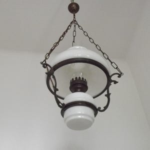 Puede incluir: Lámpara colgante antigua con una pantalla de vidrio blanco y un globo de vidrio transparente. La lámpara tiene un marco de metal oscuro y está suspendida por una cadena. Iluminación vintage.