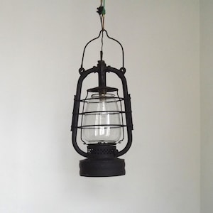 Può includere: Una lampada a sospensione a lanterna in metallo nero opaco con un globo in vetro trasparente protetto da una gabbia metallica. L'apparecchio è sospeso da un filo sottile e include una base in metallo perforato, dal design industriale.