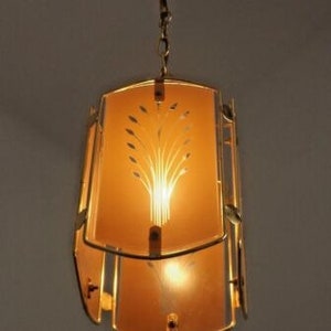 May include: A gold pendant light fixture with a frosted glass shade featuring a stylized plant design. The shade is rectangular and has a decorative metal frame.