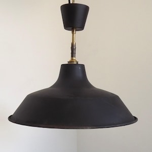 Può includere: Una lampada a sospensione in metallo nero opaco con un paralume ampio e svasato. La lampada presenta un raccordo in metallo color ottone fissato a un rosone a soffitto conico nero abbinato, sospesa tramite un cavo nero dalla parte superiore.