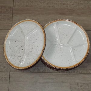 May include: A set of round ceramic plates with a white speckled finish and a brown rim. Each plate features a divided interior with five distinct sections separated by raised ridges, creating a functional layout for serving various food items.