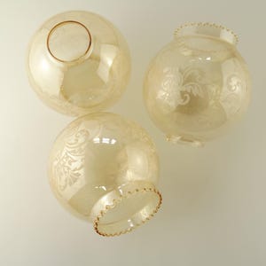 Peut inclure: Trois abat-jours sphériques en verre de couleur jaune pâle. Chaque abat-jour présente un motif floral délicat. Deux ont un bord festonné et des ouvertures en bas, tandis que le troisième a une ouverture en haut.