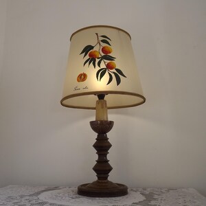 Può includere: Una lampada da tavolo in legno con un paralume a tema pesca. Il paralume è color crema con un bordo marrone e presenta un'immagine dipinta di un ramo di pesco con foglie verdi e pesche arancioni. Il testo "Frais et doux" è stampato sul paralume.