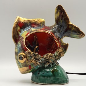 May include: A ceramic fish-shaped lamp with a vibrant color palette. The lamp features a red interior, gold accents, and a green base. The fish's exterior showcases a mix of blue, green, and gold hues, with a black power cord.