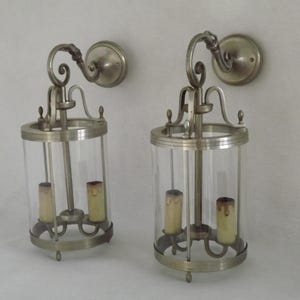 Puede incluir: Un par de apliques de pared plateadas con pantallas de vidrio transparente y dos bombillas cada una. Las apliques tienen un diseño decorativo de desplazamiento en la parte superior.