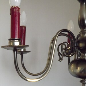 Peut inclure: Gros plan sur un lustre vintage avec une finition m&eacute;tallique. Le lustre pr&eacute;sente des bras incurv&eacute;s, des ampoules rouges en forme de bougie et des d&eacute;tails d&eacute;coratifs. Les ampoules sont blanches et les bougies sont rouge fonc&eacute;.