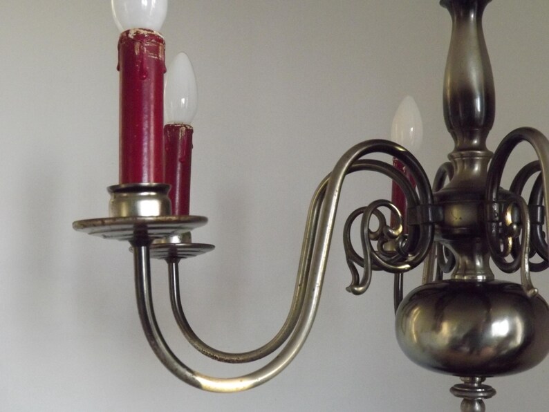 Peut inclure: Gros plan sur un lustre vintage avec une finition m&eacute;tallique. Le lustre pr&eacute;sente des bras incurv&eacute;s, des ampoules rouges en forme de bougie et des d&eacute;tails d&eacute;coratifs. Les ampoules sont blanches et les bougies sont rouge fonc&eacute;.