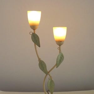 Può includere: Una lampada da tavolo decorativa con due lampade. La lampada ha una base e uno stelo color crema, con accenti di foglie verdi. Le lampade hanno la forma di fiori di tulipano ed emettono una calda luce gialla. La lampada è progettata per assomigliare a una composizione floreale.