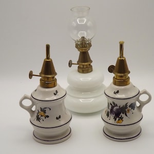 May include: Three white ceramic oil lamps with floral designs. The lamps have brass accents and glass chimneys.