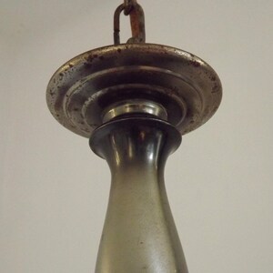 Peut inclure: Gros plan d'un luminaire vintage. Le luminaire a un auvent m&eacute;tallique circulaire terni avec un bord d&eacute;coratif. En dessous se trouve un &eacute;l&eacute;ment sombre, allong&eacute;, en forme de vase. Une cha&icirc;ne est attach&eacute;e en haut pour la suspension.