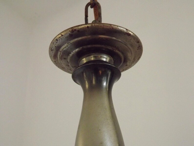 Peut inclure: Gros plan d'un luminaire vintage. Le luminaire a un auvent m&eacute;tallique circulaire terni avec un bord d&eacute;coratif. En dessous se trouve un &eacute;l&eacute;ment sombre, allong&eacute;, en forme de vase. Une cha&icirc;ne est attach&eacute;e en haut pour la suspension.