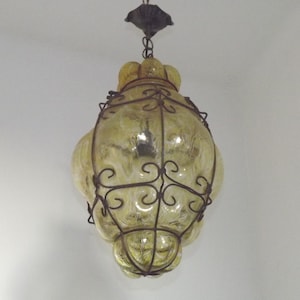 May include: A vintage hanging light fixture with a yellow glass shade and ornate dark metal frame. The glass has a textured pattern, and the fixture is suspended from a decorative ceiling mount. The overall design is reminiscent of mid-century modern lighting.