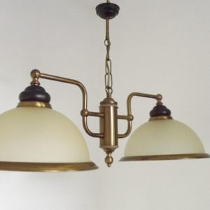 Puede incluir: Un candelabro de dos luces con acabado de latón y pantallas de vidrio color crema. La lámpara tiene un soporte central con dos brazos curvos que se extienden para sostener las pantallas. Las pantallas tienen forma de cúpula con un acento marrón oscuro.