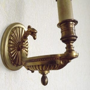 Puede incluir: Una lámpara de pared dorada con un diseño decorativo de volutas y un solo portavelas. La lámpara tiene una figura de pájaro en el lado izquierdo.