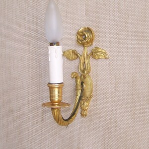 Puede incluir: Una lámpara de pared dorada con una bombilla blanca en forma de vela y un diseño decorativo de rosa.