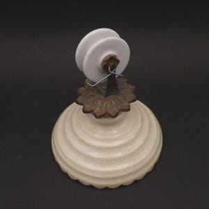 Op de afbeelding: Een vintage flesstopper met een witte keramische bovenkant en een crèmekleurige, geribbelde basis. De stopper heeft een decoratief accent van bronskleurig metaal. Het ontwerp doet denken aan antieke decoratie.