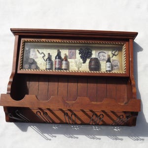 May include: A wooden wine rack with a framed display featuring miniature wine bottles, a barrel, and grapes. The rack has a shelf for bottles and metal hooks for glasses, all in a warm brown finish.