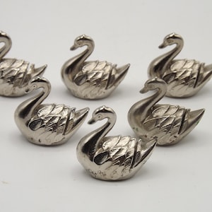 Op de afbeelding: Zes zilveren zwaanbeeldjes op een witte achtergrond. Elke zwaan heeft gedetailleerde veermotieven en een gebogen nek. Dit zijn waarschijnlijk decoratieve items of verzamelobjecten.