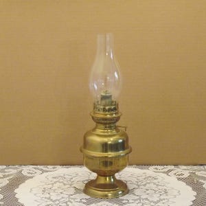 May include: A classic brass oil lamp with a clear glass chimney. The lamp features a round base, a rounded body, and detailed embellishments. It is designed to hold oil and provide illumination. The background is a neutral tone.