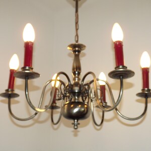 Peut inclure: Un lustre vintage avec six lumi&egrave;res en forme de bougies. Le lustre a une finition bronze avec des bras incurv&eacute;s et des bougeoirs rouges. Les ampoules sont allum&eacute;es, &eacute;mettant une lueur chaleureuse, et la fixation est suspendue &agrave; une cha&icirc;ne.