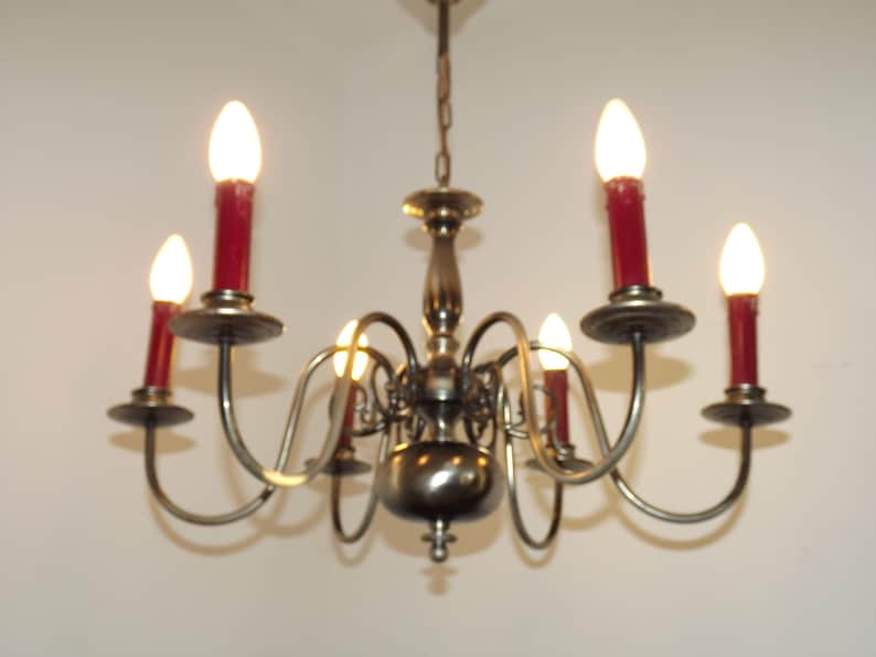 Peut inclure: Un lustre vintage avec six lumi&egrave;res en forme de bougies. Le lustre a une finition bronze avec des bras incurv&eacute;s et des bougeoirs rouges. Les ampoules sont allum&eacute;es, &eacute;mettant une lueur chaleureuse, et la fixation est suspendue &agrave; une cha&icirc;ne.