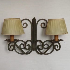 Puede incluir: Un aplique de pared doble con un marco de metal oscuro con volutas intrincadas y dos pantallas de tela plisada de color beige. La lámpara tiene una placa de montaje vertical central y dos brazos que se extienden hacia afuera.