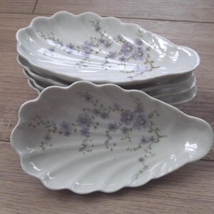 Könnte beinhalten: Ein Stapel weißer Porzellanschalen in Muschelform mit gewelltem Rand. Jede Schale ist mit einem zarten Blumenmuster aus kleinen hellvioletten Blüten und dünnen braunen Stielen verziert, die sich wie ein Zweig über die Mitte erstrecken.
