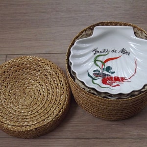 May include: A white ceramic dish shaped like a seashell with a red shrimp and green seaweed design. The dish says "Fruits de Mer" in black lettering. The dish is inside a woven basket.