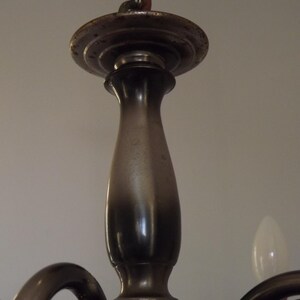 Peut inclure: Gros plan sur la colonne centrale et la plaque de montage d'un lustre vintage. Le luminaire en m&eacute;tal pr&eacute;sente une finition sombre et vieillie, avec une ampoule visible de chaque c&ocirc;t&eacute;. Le design sugg&egrave;re un style ancien ou r&eacute;tro.