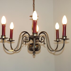 Peut inclure: Un lustre vintage avec six lumi&egrave;res en forme de bougie. Le lustre a une monture en m&eacute;tal fonc&eacute; avec des bras incurv&eacute;s et des volutes d&eacute;coratives. Les bougeoirs sont surmont&eacute;s d'ampoules rouges en forme de bougie, et les lumi&egrave;res sont allum&eacute;es.