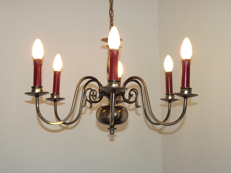 Peut inclure: Un lustre vintage avec six lumi&egrave;res en forme de bougie. Le lustre a une monture en m&eacute;tal fonc&eacute; avec des bras incurv&eacute;s et des volutes d&eacute;coratives. Les bougeoirs sont surmont&eacute;s d'ampoules rouges en forme de bougie, et les lumi&egrave;res sont allum&eacute;es.