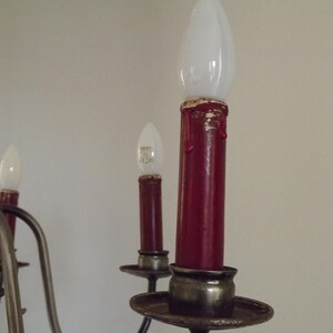 Peut inclure: Gros plan sur un lustre avec trois lumi&egrave;res rouges en forme de bougie. Les lumi&egrave;res ont des ampoules blanches en forme de flamme. Le lustre a une armature en m&eacute;tal fonc&eacute; avec des courbes d&eacute;coratives et une base ronde. Le fond est un mur uni, blanc cass&eacute;.