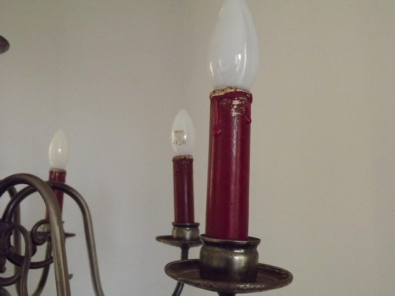 Peut inclure: Gros plan sur un lustre avec trois lumi&egrave;res rouges en forme de bougie. Les lumi&egrave;res ont des ampoules blanches en forme de flamme. Le lustre a une armature en m&eacute;tal fonc&eacute; avec des courbes d&eacute;coratives et une base ronde. Le fond est un mur uni, blanc cass&eacute;.