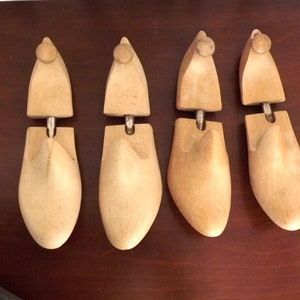 May include: Four wooden shoe trees, each with a metal bar in the middle, are arranged in a row on a brown surface. The shoe trees are light brown and have a smooth finish.