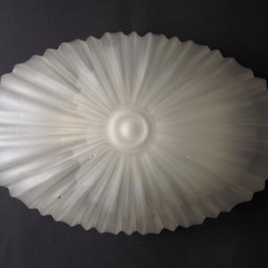 Könnte beinhalten: Eine ovale Leuchte aus mattiertem Glas mit radialem, geriffeltem Design. Die Mitte weist ein kleines, kreisförmiges Detail auf. Die Kanten sind gewellt. Die Leuchte hat eine weiche, weiße Farbe.