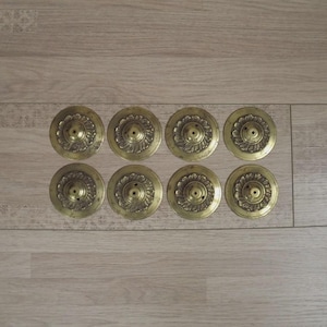 Könnte beinhalten: Acht antike Messing-Räuchergefäßdeckel mit floralen Designs. Jeder Deckel ist kreisförmig mit einem zentralen Loch und dekorativen Details. Die Deckel sind in zwei Reihen zu je vier Stück auf einer hellen Holzoberfläche angeordnet.