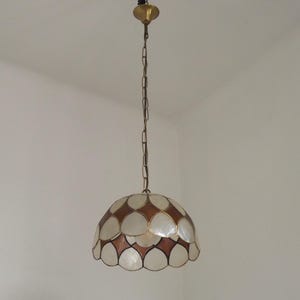 Peut inclure: Une lampe suspendue dotée d'un abat-jour en forme de dôme composé de pièces blanches superposées rappelant des coquillages et d'accents bruns en forme de losange. Le luminaire est suspendu par une chaîne en métal doré avec une rosace assortie.